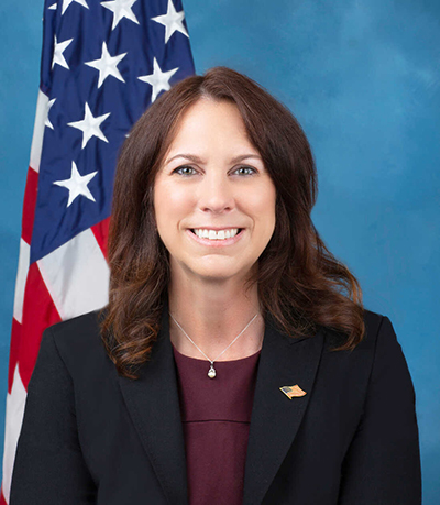 A woman with dark hair in a dark jacket and wearing an American flag on her lapel, stands in front of an American flag.
