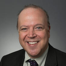 Smiling, gray-haired man wearing a black suit and tie.