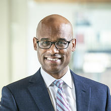 Smiling man with black glasses wearing dark suit and striped tie.